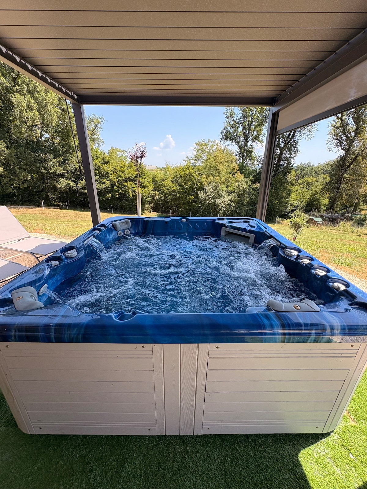 A hot tub is situated under a pergola, surrounded by a lush green garden. Bubbles can be seen rising from the water, creating a soothing atmosphere. Sunlight filters through the trees in the background, enhancing the outdoor tranquility of the space.