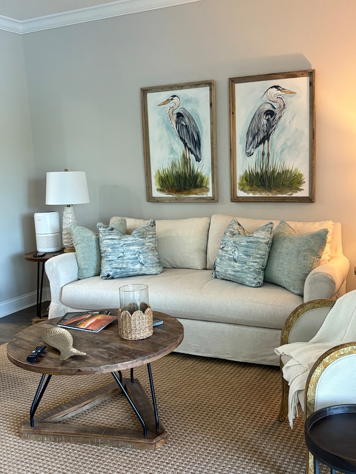 A comfortable seating area is presented with a light-colored sofa adorned with textured pillows. Two framed paintings of herons adorn the wall. A wooden coffee table displays a decorative item and a magazine, with a lamp providing soft illumination in the corner.