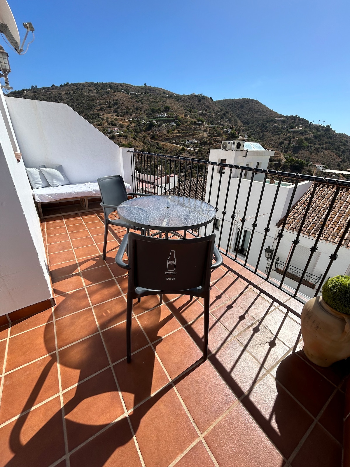 A sunlit terrace is shown, featuring a round glass table surrounded by four chairs. A comfortable lounge area is positioned against a wall, with decorative pillows arranged neatly. The backdrop presents scenic views of the surrounding hills, creating a serene outdoor space.