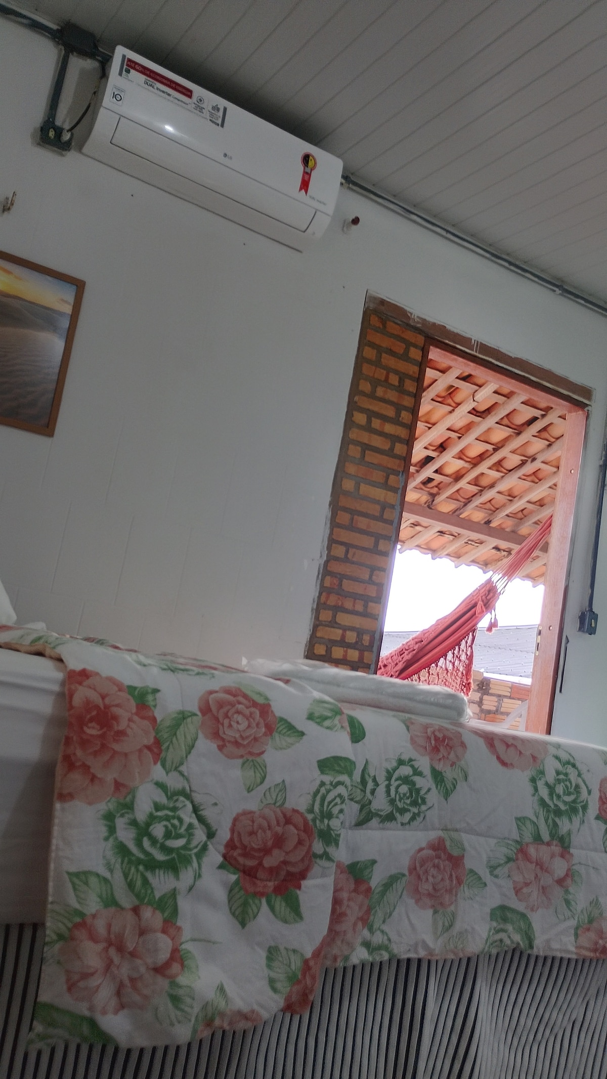 A cozy sleeping area is presented, featuring a floral-patterned blanket draped over a spacious bed. A window reveals an outdoor space with a hammock, allowing natural light to filter in. A small framed picture is visible on the wall, enhancing the room's decor.