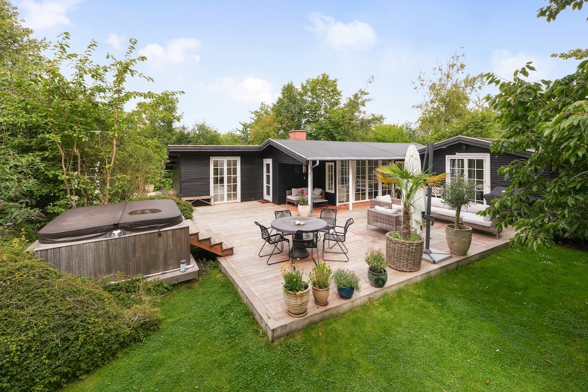 A charming summer house is surrounded by lush greenery and features a spacious wooden deck. Outdoor seating arrangements can be seen alongside a jacuzzi, inviting relaxation amidst nature. Potted plants add a touch of greenery, creating a serene outdoor space.