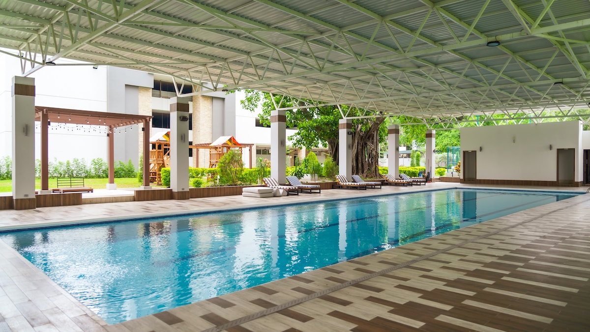An indoor swimming pool is showcased with crystal-clear water reflecting the surrounding light. Comfortable lounge chairs are arranged neatly along the poolside. Large windows allow natural light to fill the space, connecting it to the greenery visible outside.