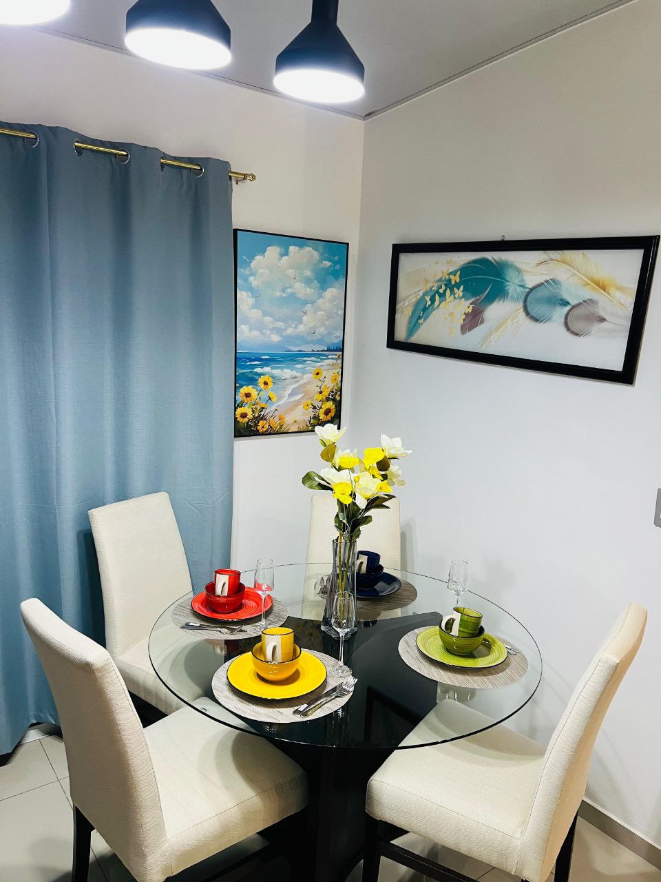 A round glass dining table is set for four, featuring colorful plates and drinks. A vase of fresh flowers sits in the center, with a painting of a beach scene visible on the wall. Soft blue curtains frame the window, enhancing the cozy dining area.