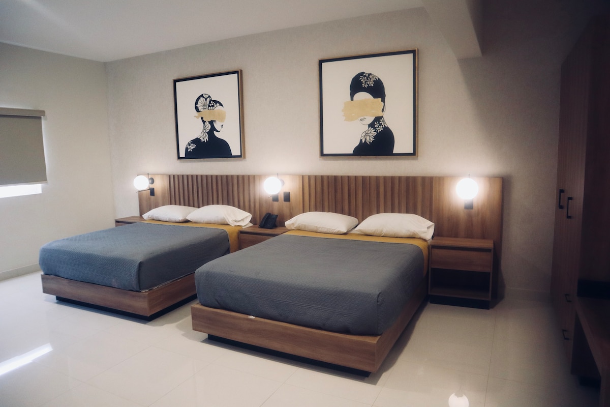 A bright and airy guest room is presented with two spacious beds, each adorned with minimalistic grey linens. Above the beds, two framed artworks depict figures with subtle details. Soft lighting is provided by modern bedside lamps on stylish wooden headboards.