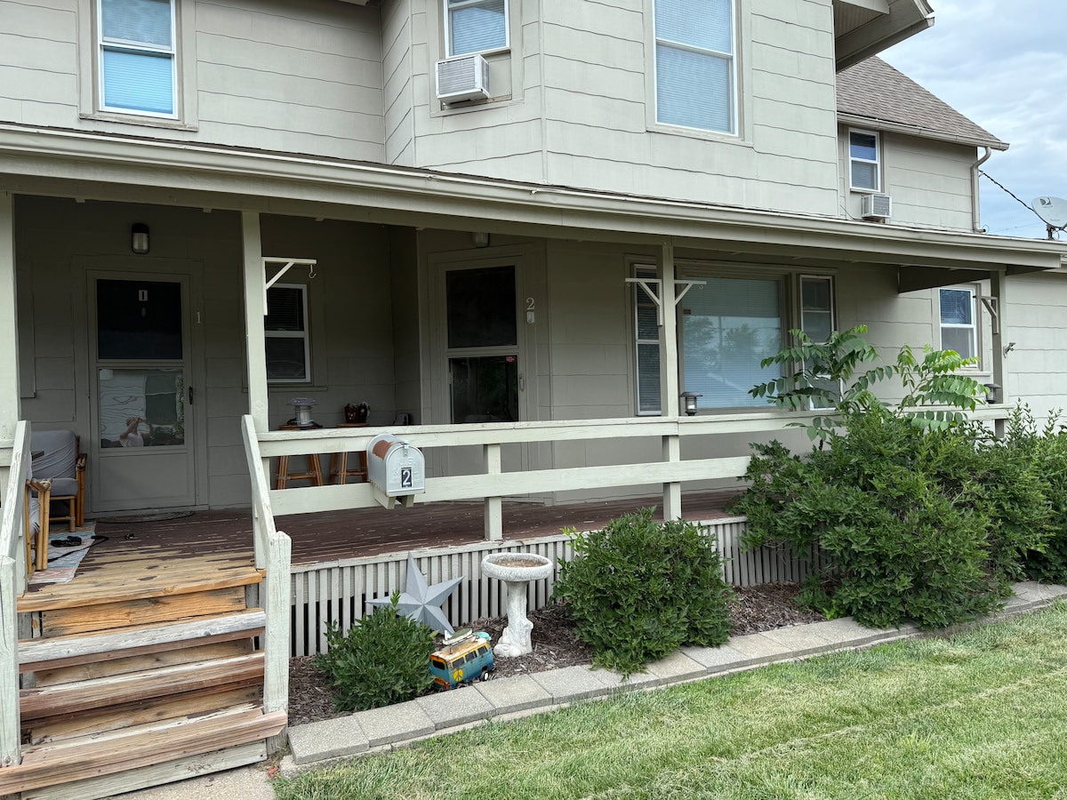 An inviting exterior porch is visible, featuring wooden steps leading up to a light grey entrance door. Green shrubs and a small area of lawn surround the home, while a seating arrangement can be seen on the porch. Windows allow natural light to fill the space.
