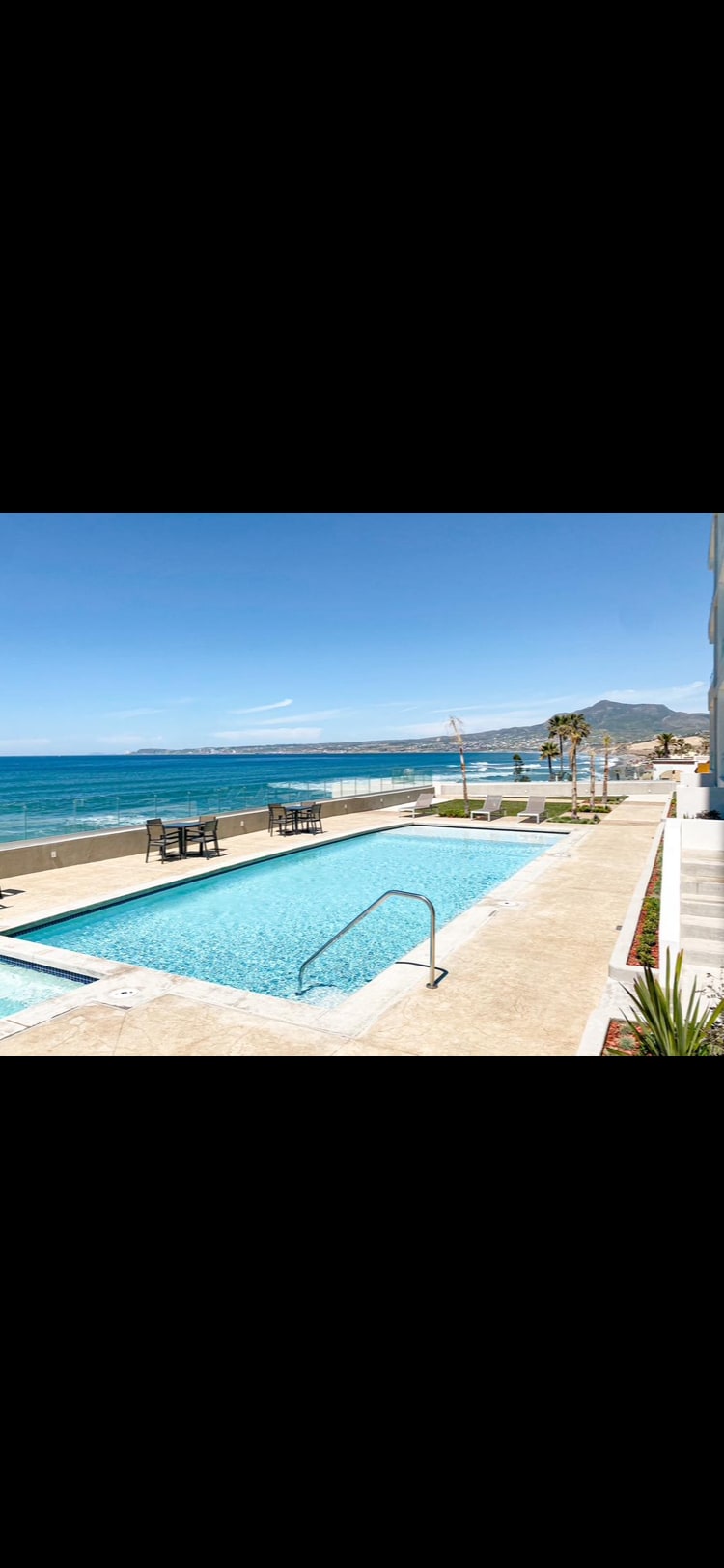 A clear blue pool is positioned alongside a sandy beach, with ocean waves visible in the distance. Lounge chairs face the water, while palm trees line the shore. The inviting atmosphere is enhanced by the sunshine illuminating the area, creating a serene outdoor space.