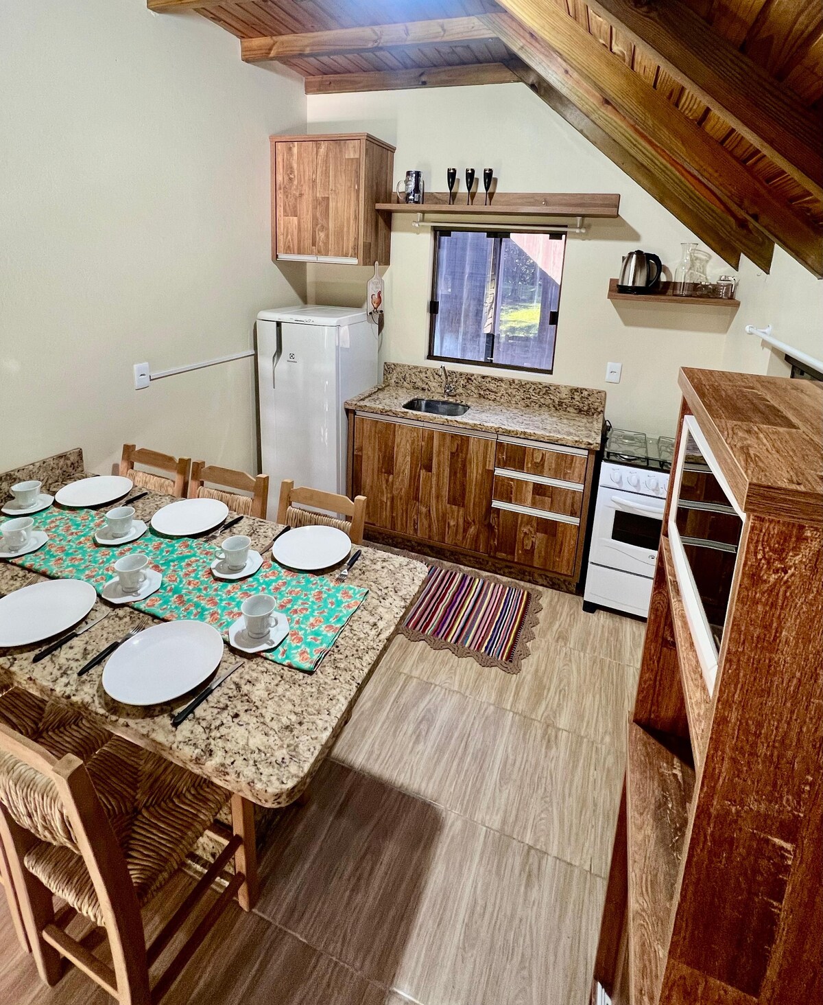 A well-equipped kitchen area features wooden cabinetry and granite countertops. A dining table is set for several guests, with plates and cups arranged neatly. Appliances include a refrigerator, stove, and kettle, while natural light filters through a nearby window, highlighting the space.