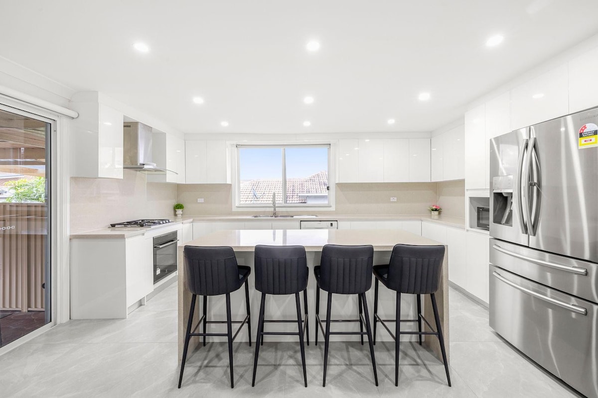 A modern kitchen features sleek white cabinetry and a large island with seating for four. Stainless steel appliances are visible, including a refrigerator and an oven. Natural light enters through a sizable window above the sink, enhancing the clean and spacious layout.