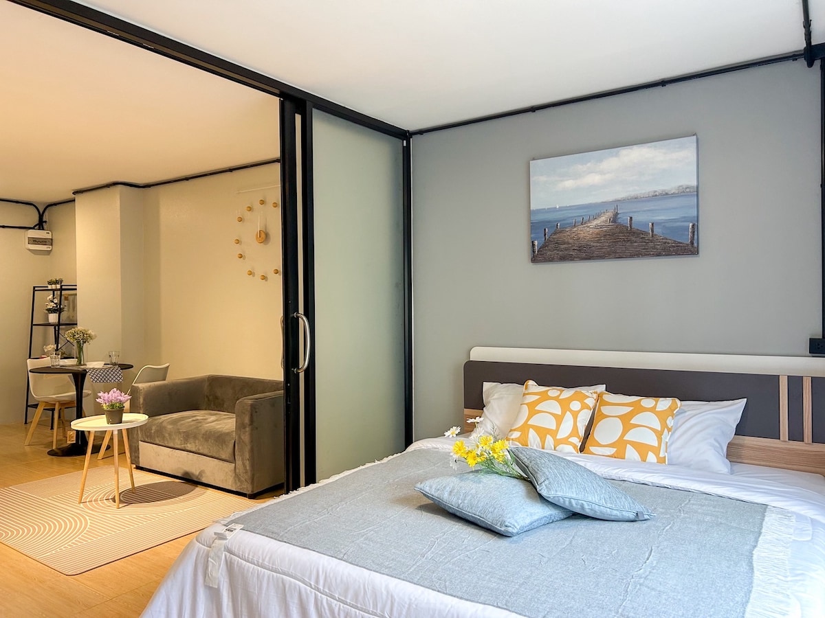 A well-lit sleeping area features a spacious bed adorned with light linens and decorative pillows. A painting of a pier hangs above the bed. In the background, a cozy seating area is visible, with a gray couch and a small dining table set beside it.