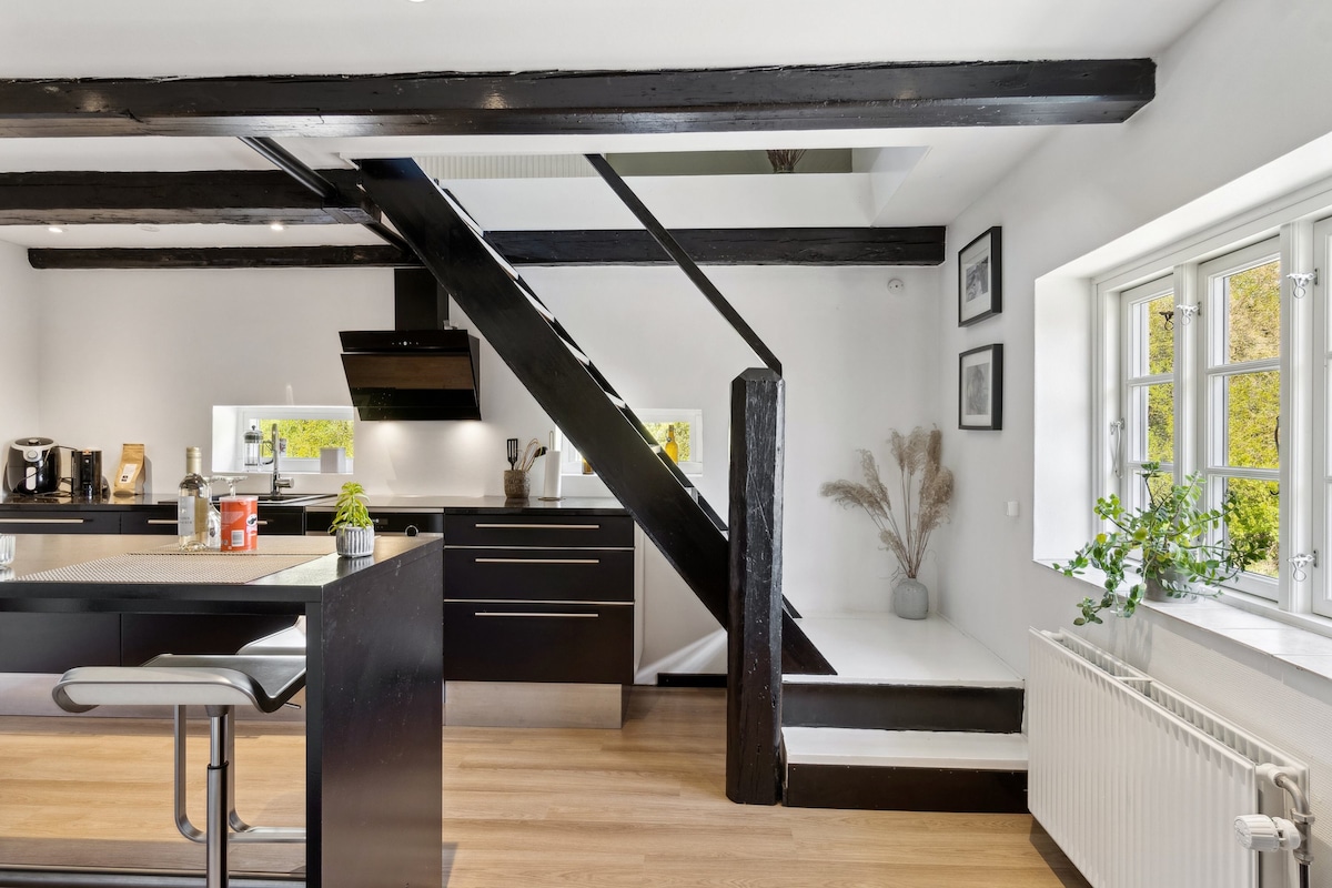 A modern kitchen layout is showcased with dark cabinetry and sleek countertops. Exposed wooden beams add character along with a staircase leading to the upper level. Natural light fills the space through large windows, complemented by decorative plants positioned nearby.