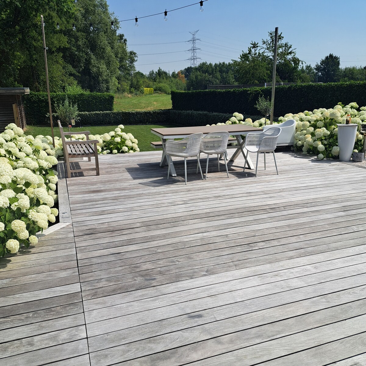 An outdoor deck features a wooden surface, surrounded by lush greenery and clusters of white flowers. A dining table with four chairs is positioned beside a compact seating area, providing a space for relaxation. Soft sunlight highlights the inviting outdoor setting.