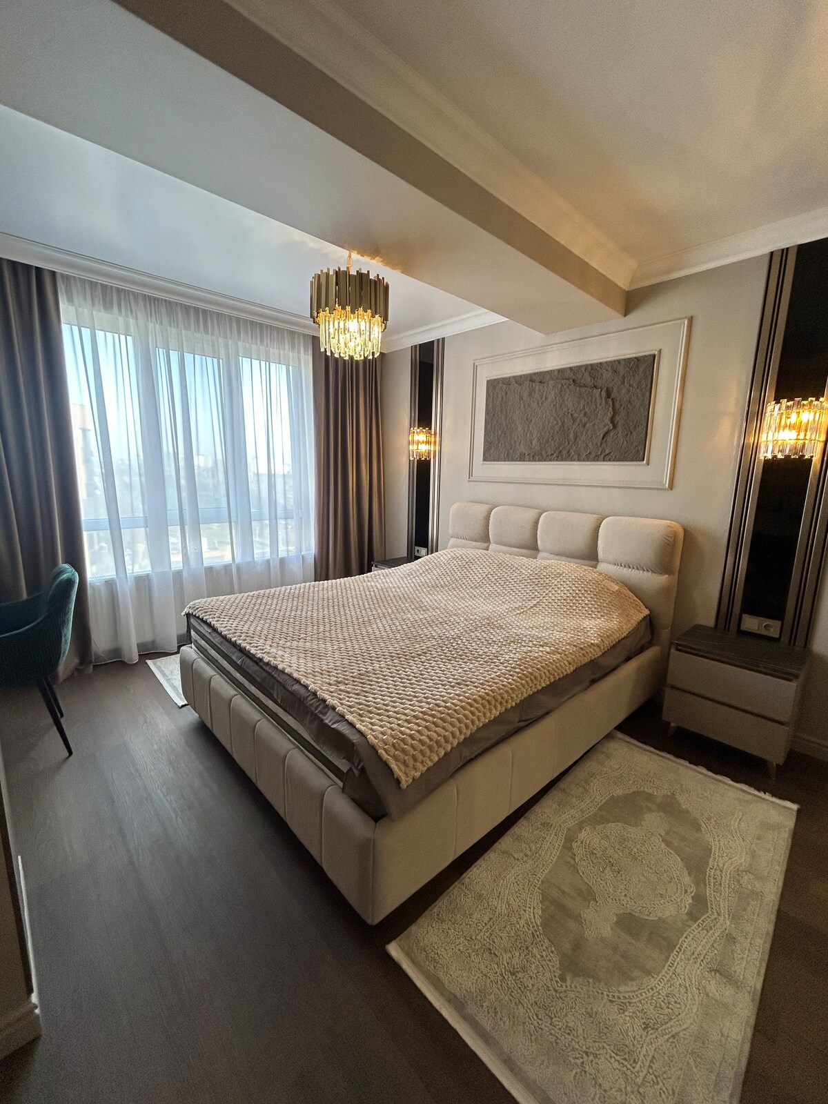 A well-appointed bedroom features a spacious bed with a neutral-toned blanket. Natural light floods the room through large floor-to-ceiling windows, and modern lighting accents are positioned on either side of the bed. A small writing desk and a plush green chair are visible in the corner.