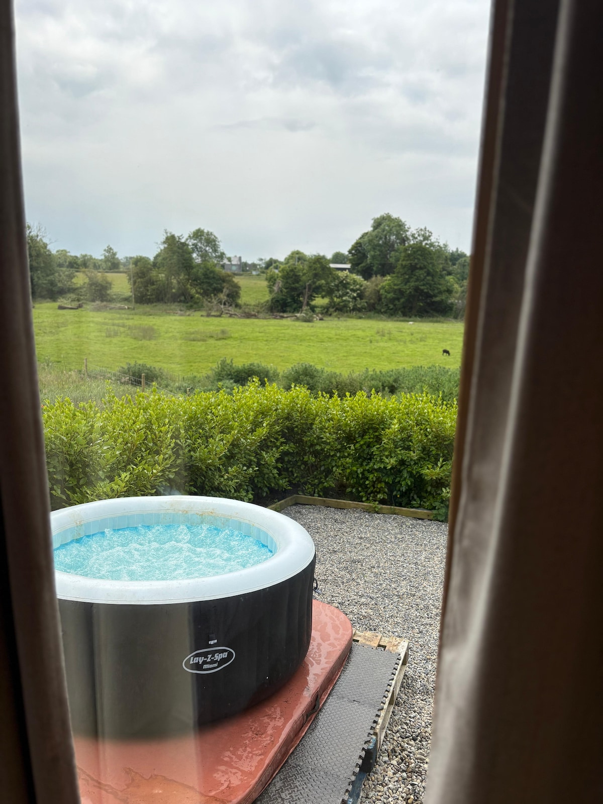 A private hot tub is viewed through a window, set on a patio overlooking a lush green landscape. The clear water of the tub contrasts with the surrounding greenery, creating a serene outdoor retreat. The scene is framed by sheer curtains for added privacy.