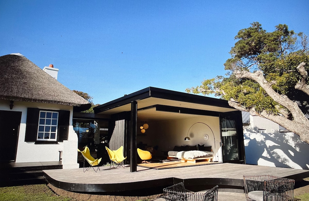 A modern cottage with a thatched roof is complemented by a spacious wooden deck. Two bright yellow chairs sit outside, inviting relaxation, while large glass walls provide a view into the contemporary interior, showcasing a cozy sleeping area.