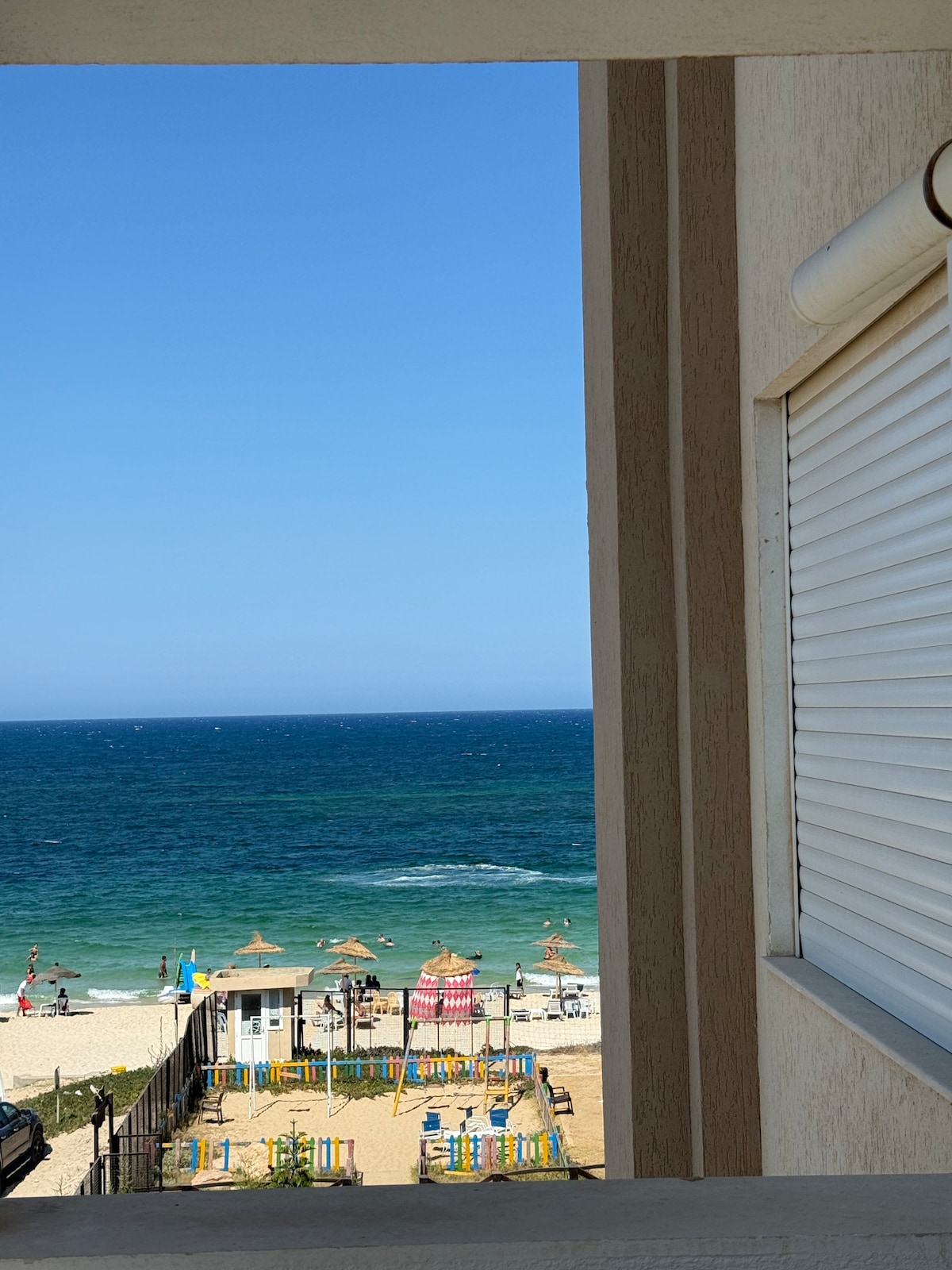 A view is captured through a window framing the vibrant beach below, where colorful umbrellas and loungers are positioned along the shoreline. The deep blue ocean stretches into the horizon, gently lapping against the sand, with a clear blue sky overhead.