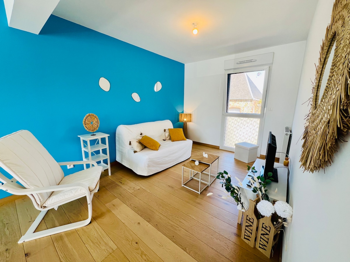 A bright living area features a white sofa with decorative pillows, a glass coffee table, and a side table. A comfortable white armchair is present, alongside a television. A large window allows natural light to fill the space, complemented by a blue accent wall and wooden flooring.