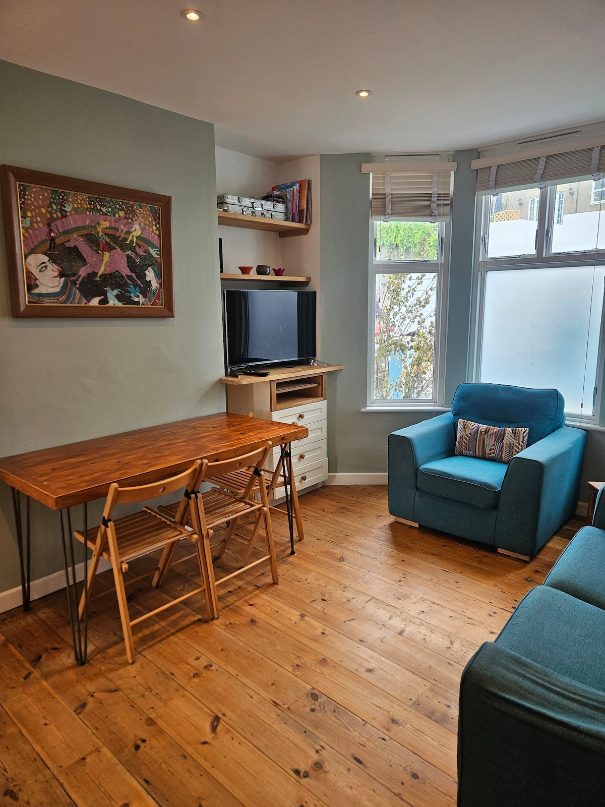 A cozy living area features a blue armchair and a sofa facing a compact wooden dining table with folding chairs. Natural light filters through window panes adorned with blinds, illuminating the polished wooden floor. A vibrant artwork decorates the wall, adding a touch of color.