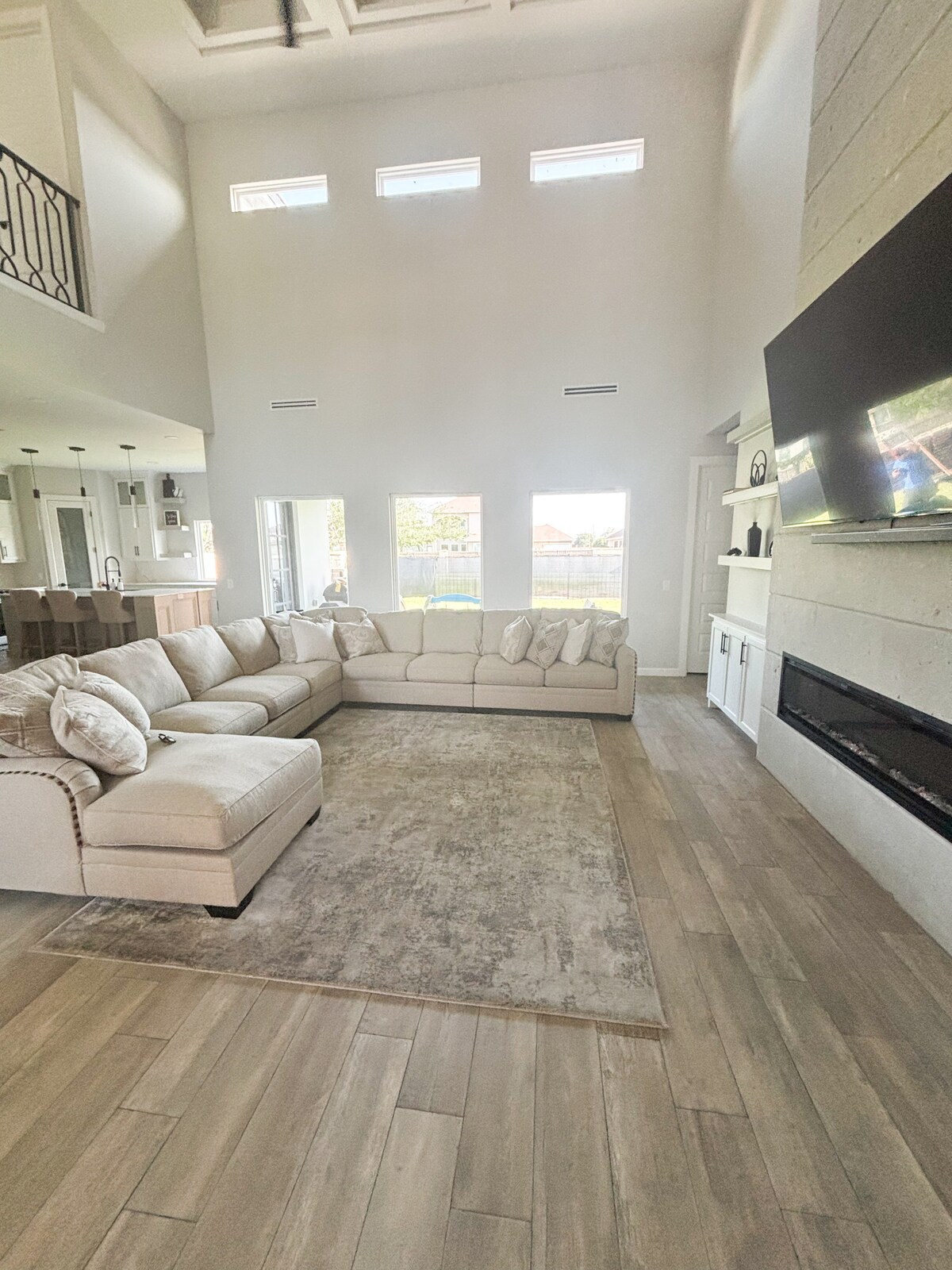 A spacious living area features a large sectional sofa arranged around a neutral area rug. Natural light enters through several windows, illuminating the room's high ceilings and modern fireplace beneath a mounted television.