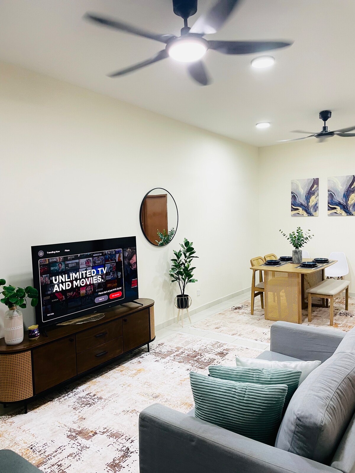 The living area features a large flat-screen TV displaying a streaming service. A round mirror hangs above a wooden side table, while a cozy sofa adorned with decorative cushions faces the screen. A small dining table is set nearby, complemented by indoor plants and soft lighting.