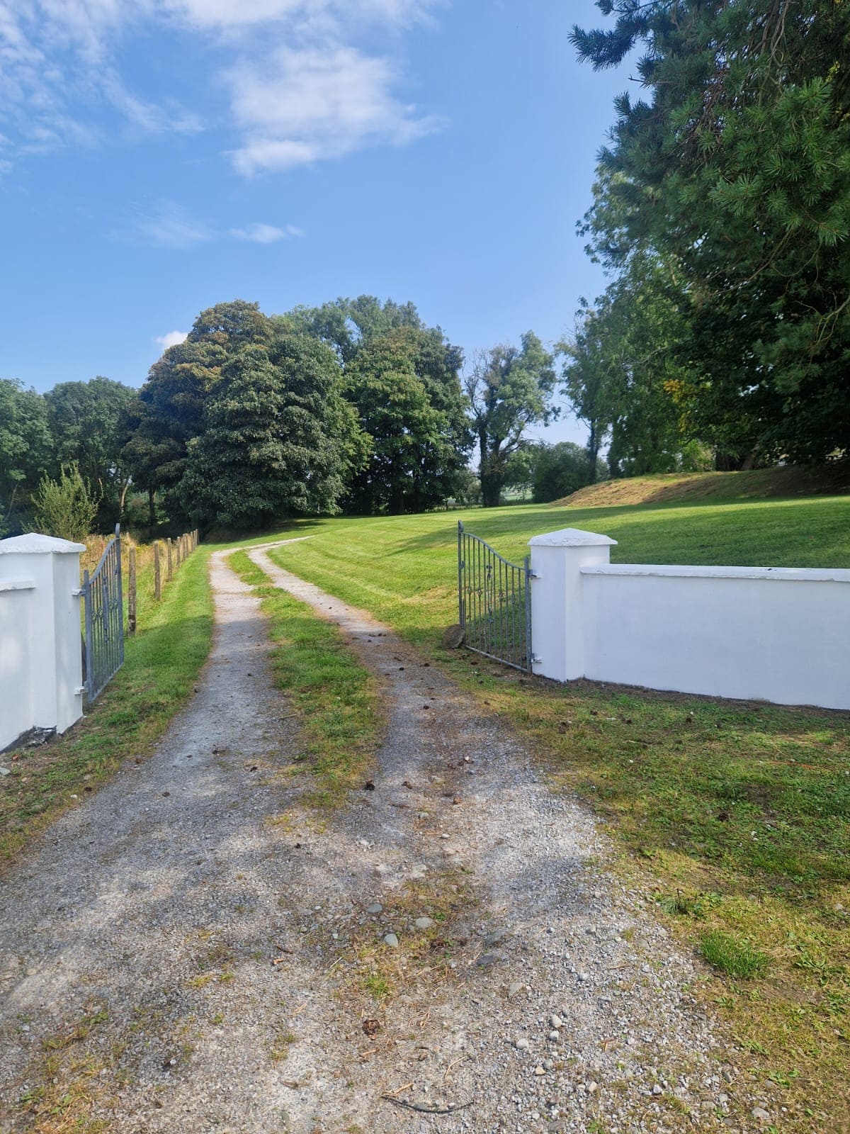 A gravel driveway meanders through a lush green landscape, bordered by tall trees. The entrance features a white gate that opens to a well-maintained garden area, enhancing the inviting nature of the farmhouse's surroundings.
