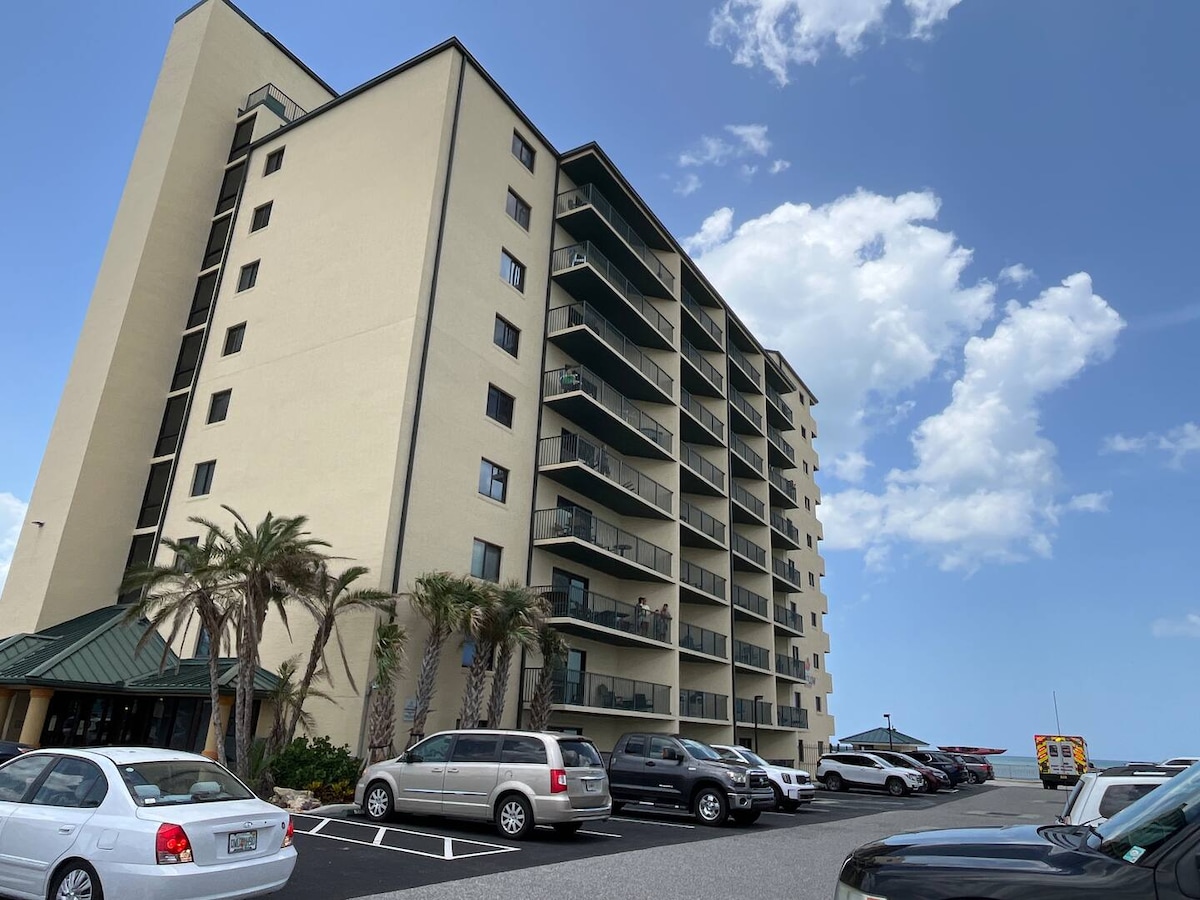 The image captures a multi-story building with a pale exterior, surrounded by palm trees and a parking area. The structure includes balconies on several floors, and the nearby beach is visible in the background, highlighting the condo's proximity to the ocean.