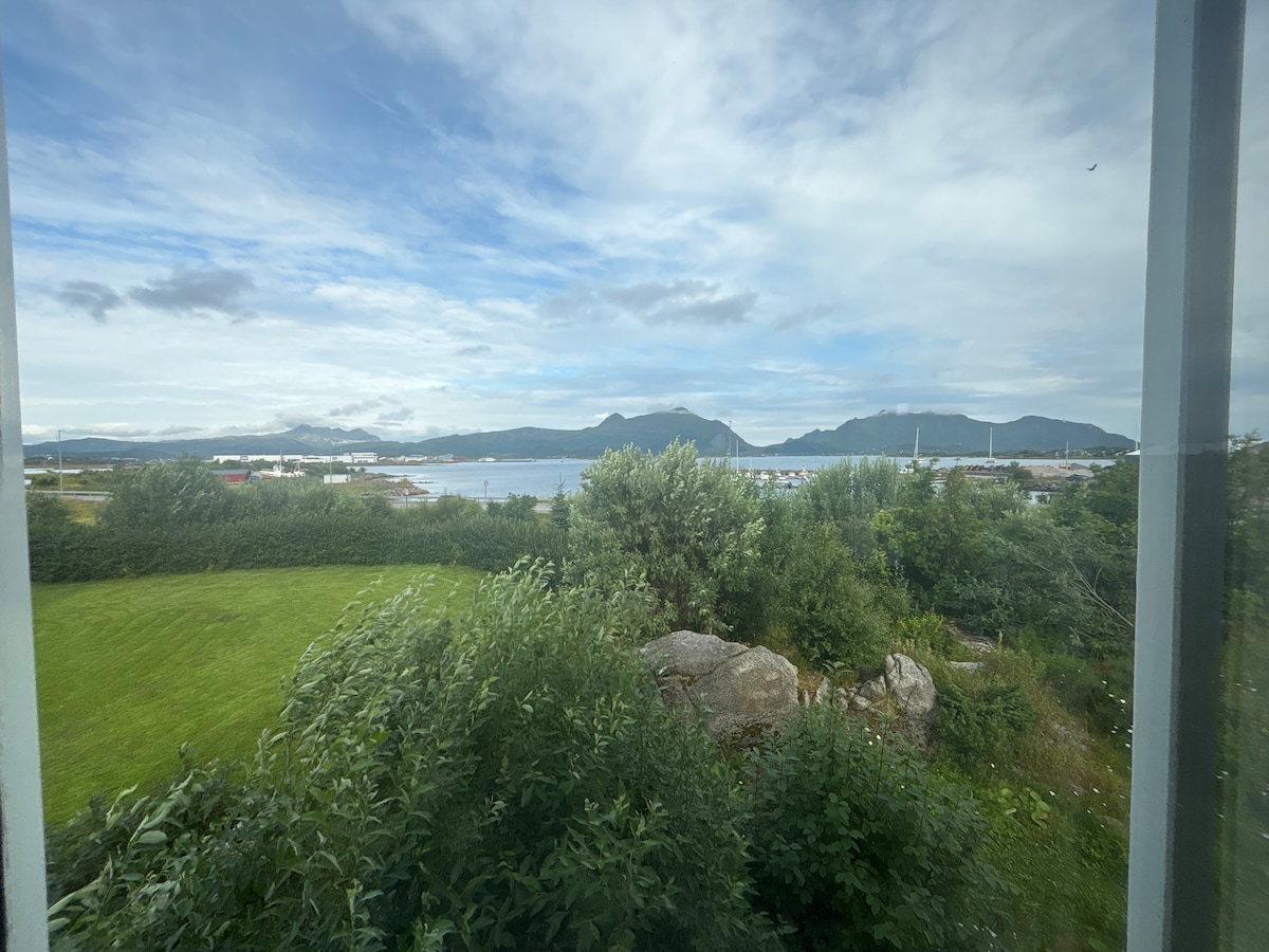 A view from a window showcases a tranquil landscape featuring lush greenery in the foreground. A glimpse of the water is seen beyond, with hills rising in the background under a cloudy sky. The scene captures a serene coastal setting.
