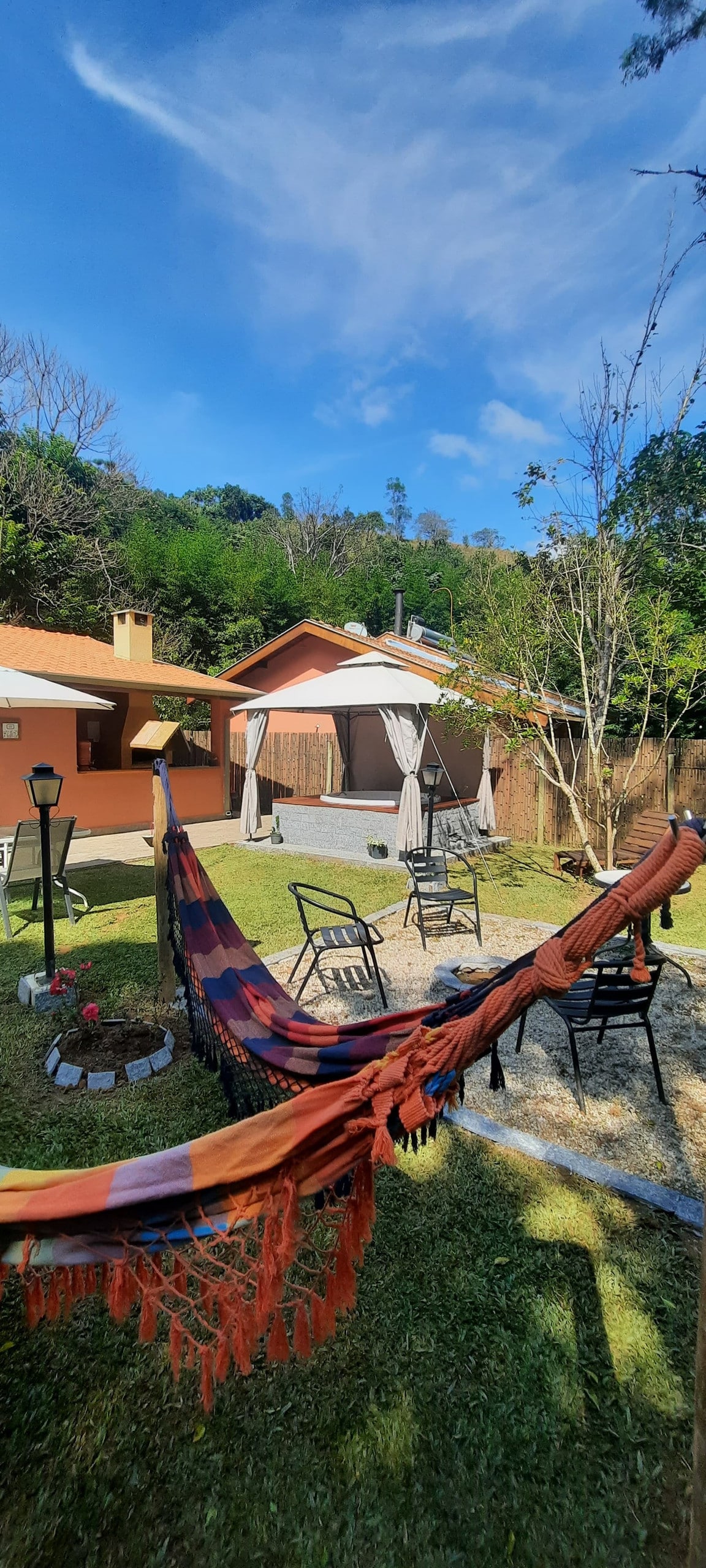 A relaxing outdoor space features a colorful hammock draped between trees, surrounded by a garden. Several chairs are arranged around a circular stone fire pit. In the background, two houses are visible under a clear blue sky, with greenery enhancing the serene atmosphere.
