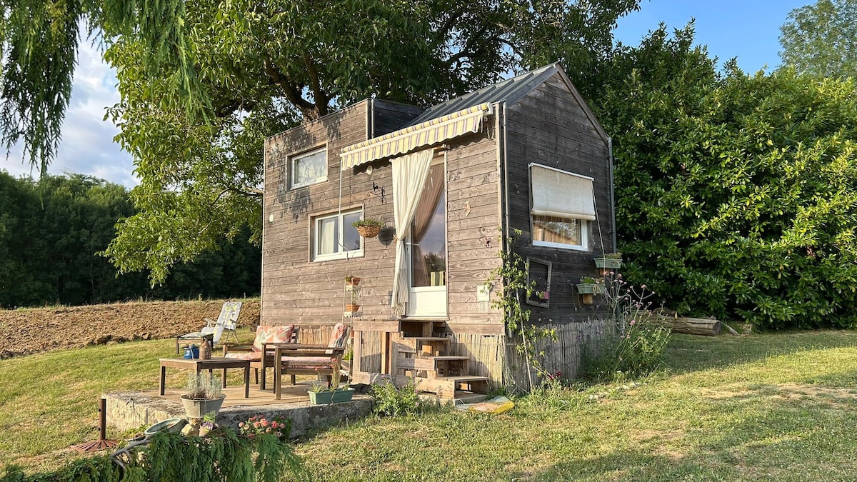 A charming tiny house stands amidst greenery, featuring a small private terrace with outdoor seating. The structure is adorned with plants and a cozy awning, while large windows allow natural light to fill the interior. Surrounding gardens and a nearby tree offer additional shade.