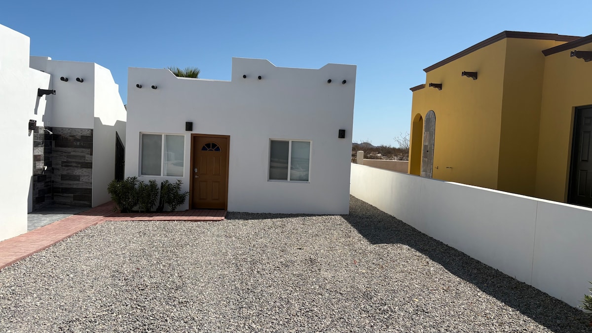 A charming white casita is positioned with a distinctive brown door. The surrounding area is paved with gravel, and two buildings, one in a bright yellow hue, can be seen in the background under a clear blue sky.