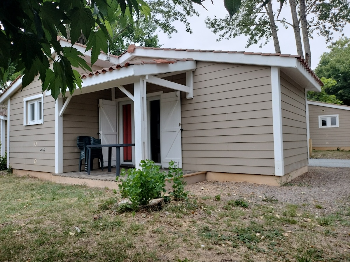 A single-story home is depicted with a light brown exterior, featuring a covered porch supported by white beams. A small table with chairs is positioned on the porch, and a door on the right side leads into the interior. Nearby green foliage adds to the natural setting.