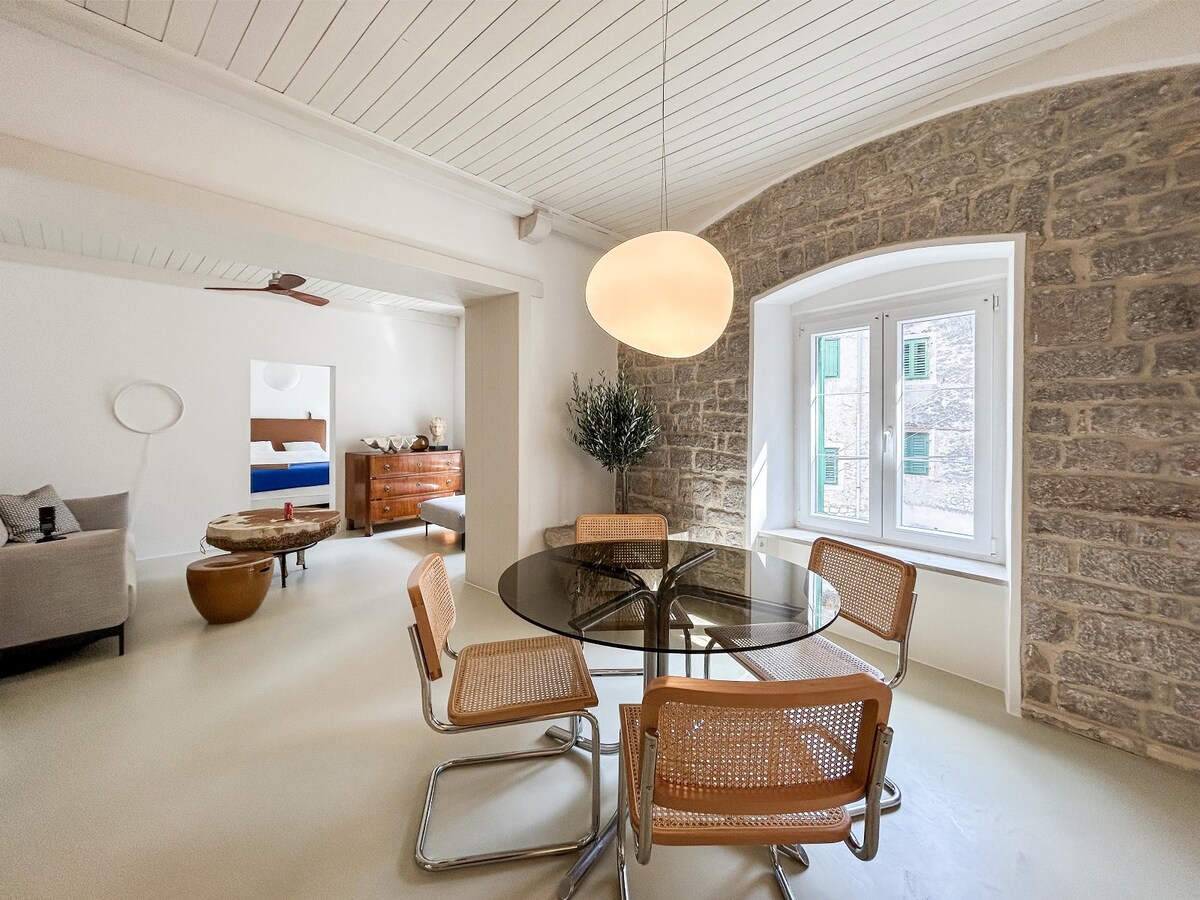 A dining area features a round glass table surrounded by four woven chairs. Soft natural light filters through two south-facing windows, illuminating the space. A stone wall provides a textured backdrop, enhancing the room's character, while a ceiling fixture offers gentle lighting.