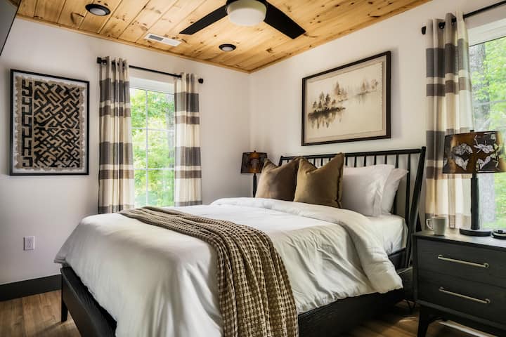 Un havre de paix douillet et moderne en montagne pour une nuit de repos parfaite. Cette chambre d'invités lumineuse est équipée d'un lit queen moelleux, d'œuvres d'art locales uniques et d'un plafond chaleureux en planches de bois avec un ventilateur rafraîchissant. De grandes fenêtres offrent une vue paisible sur les arbres environnants.