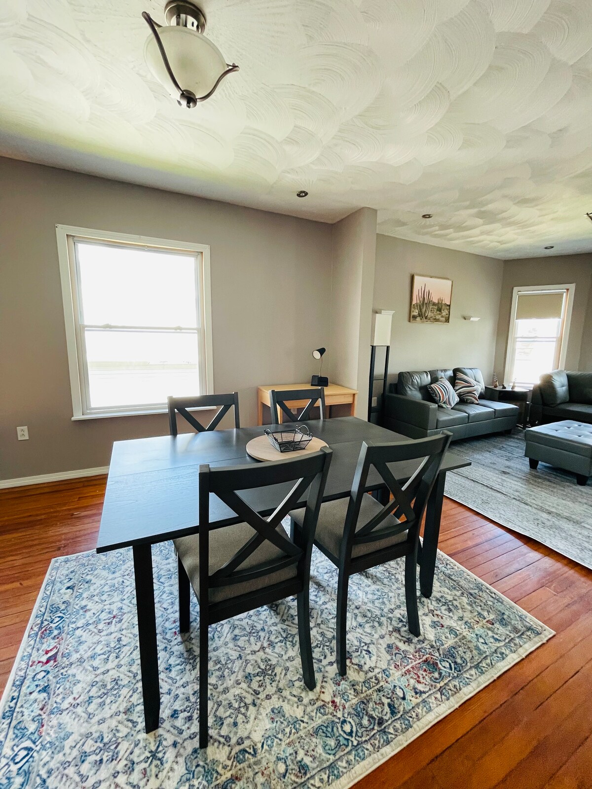A dining area is shown with a dark wooden table surrounded by four chairs. A simple decorative plate is placed on the table. Natural light enters through a nearby window, illuminating the space and highlighting a soft area rug beneath the table.