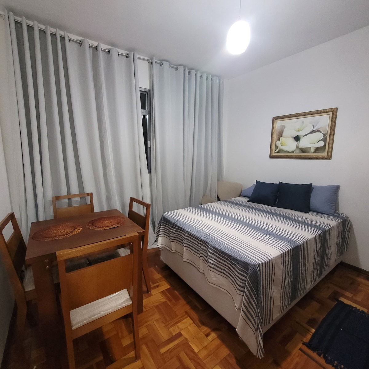 A spacious sleeping area features a comfortable bed dressed in a striped blanket, complemented by several accent pillows. A wooden dining table with four chairs is visible nearby, and light drapes softly filter through sheer curtains. A wall painting adds a touch of decor.