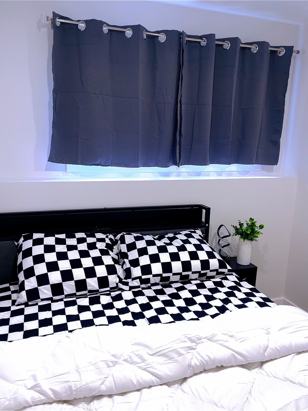A modern sleeping area features a queen-sized bed with a black and white checkered duvet and two pillows. A bedside table is positioned next to the bed, holding a small plant. Light curtains cover a window, providing a gentle diffusion of natural light.