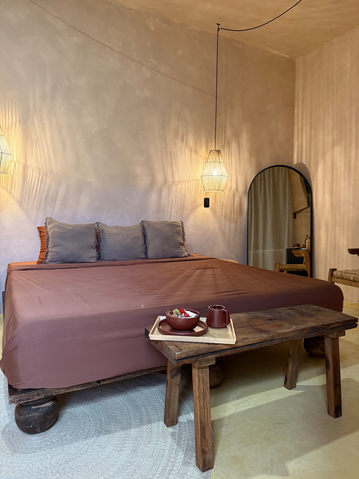 A spacious bed with a brown coverlet is paired with soft gray pillows. A wooden table holds a tray with a bowl, and an arched mirror reflects the room's warm tones. Soft light is provided by decorative hanging fixtures.