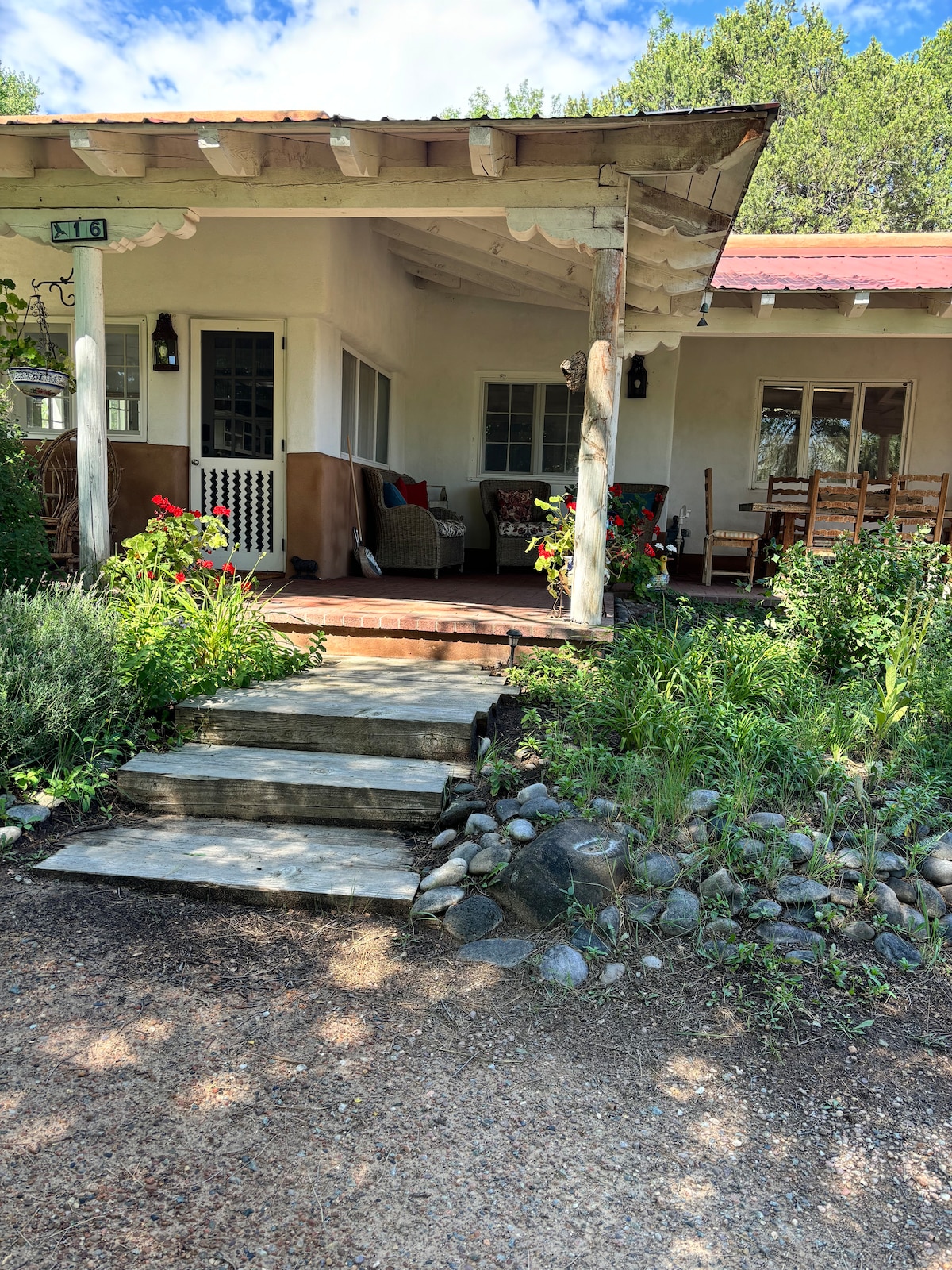 The front of the Country House features a covered porch with inviting seating areas adorned with colorful flowers. A stone pathway leads to the entrance, which is flanked by lush greenery and a blend of shrubs and rocks, creating a serene outdoor environment.