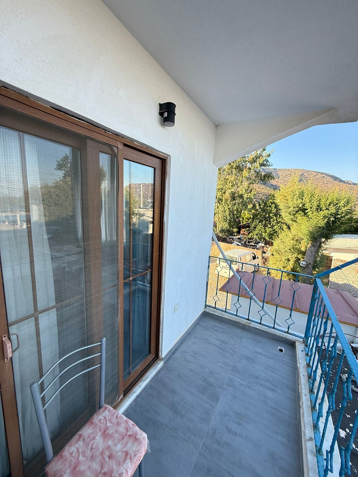 A balcony space is presented, featuring a light-colored floor and a small metal chair. Floor-to-ceiling glass doors offer a view of the outdoors, with greenery and hills visible in the background. A light fixture is positioned near the ceiling.