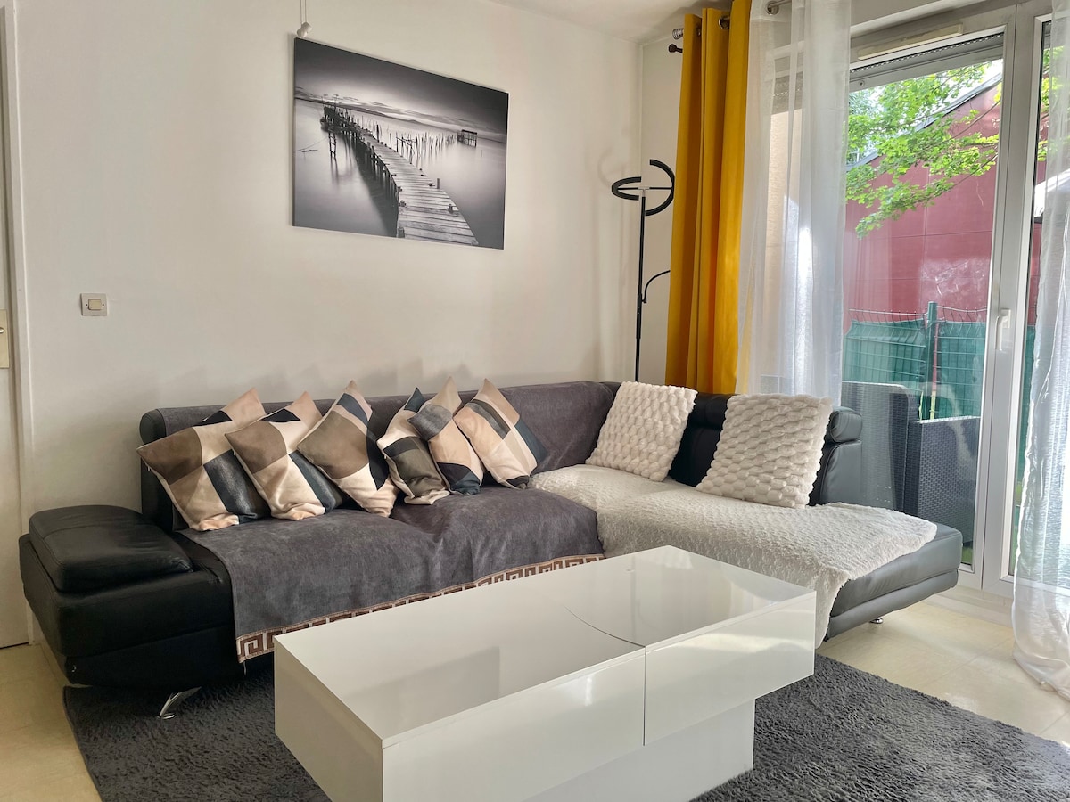A modern living area features a sectional sofa adorned with various decorative cushions. A sleek white coffee table is positioned in front of the sofa. Bright curtains and large windows allow natural light to illuminate the space, enhancing its welcoming atmosphere.