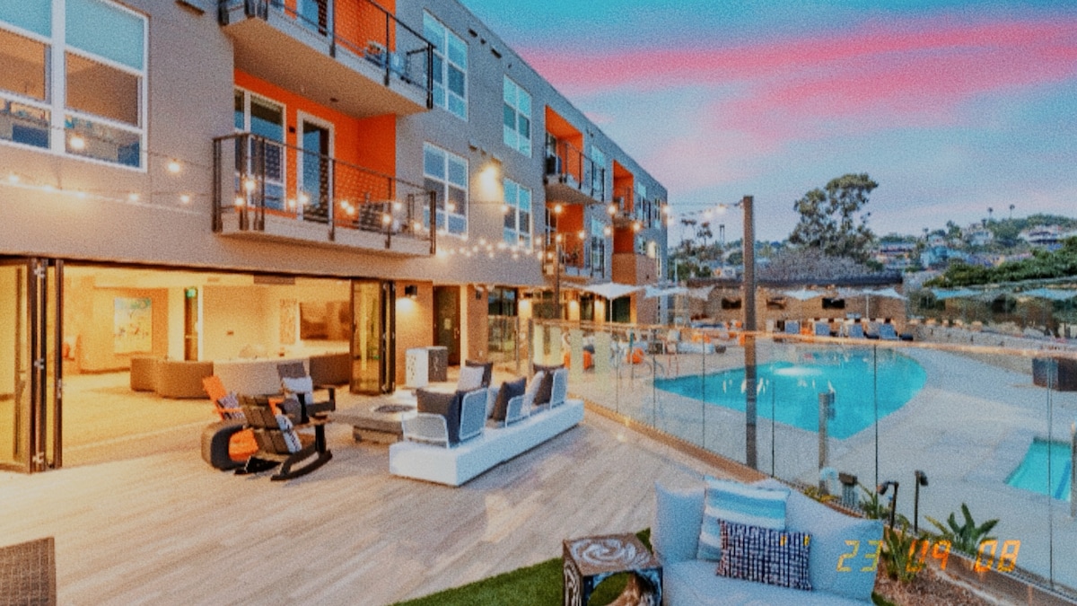 Outdoor seating surrounds a modern pool area, enhanced by soft lighting from string lights above. A spacious deck accommodates lounge chairs and a table, offering a view of the water feature and well-maintained greenery in the background.