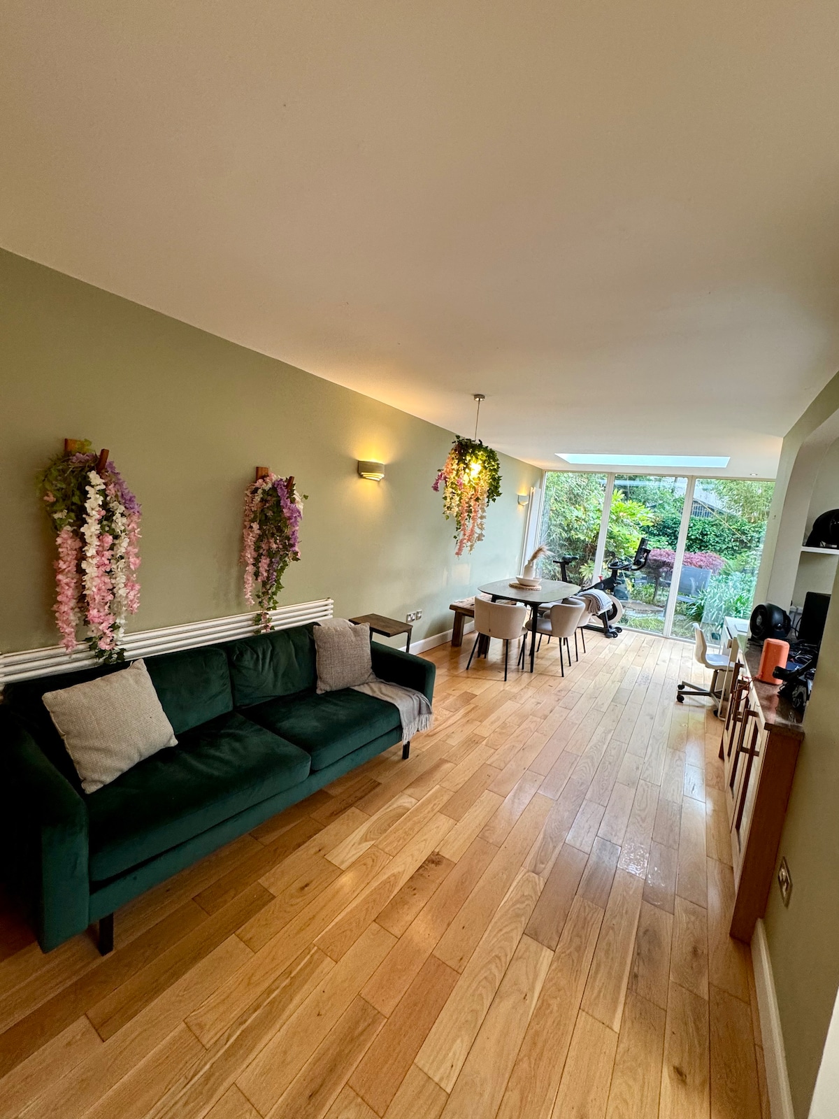 A cozy living area features a green sofa with soft cushions, complemented by decorative flower arrangements hanging on the walls. A dining table with chairs is visible, and large glass doors allow natural light to illuminate the room and provide a view of the garden.