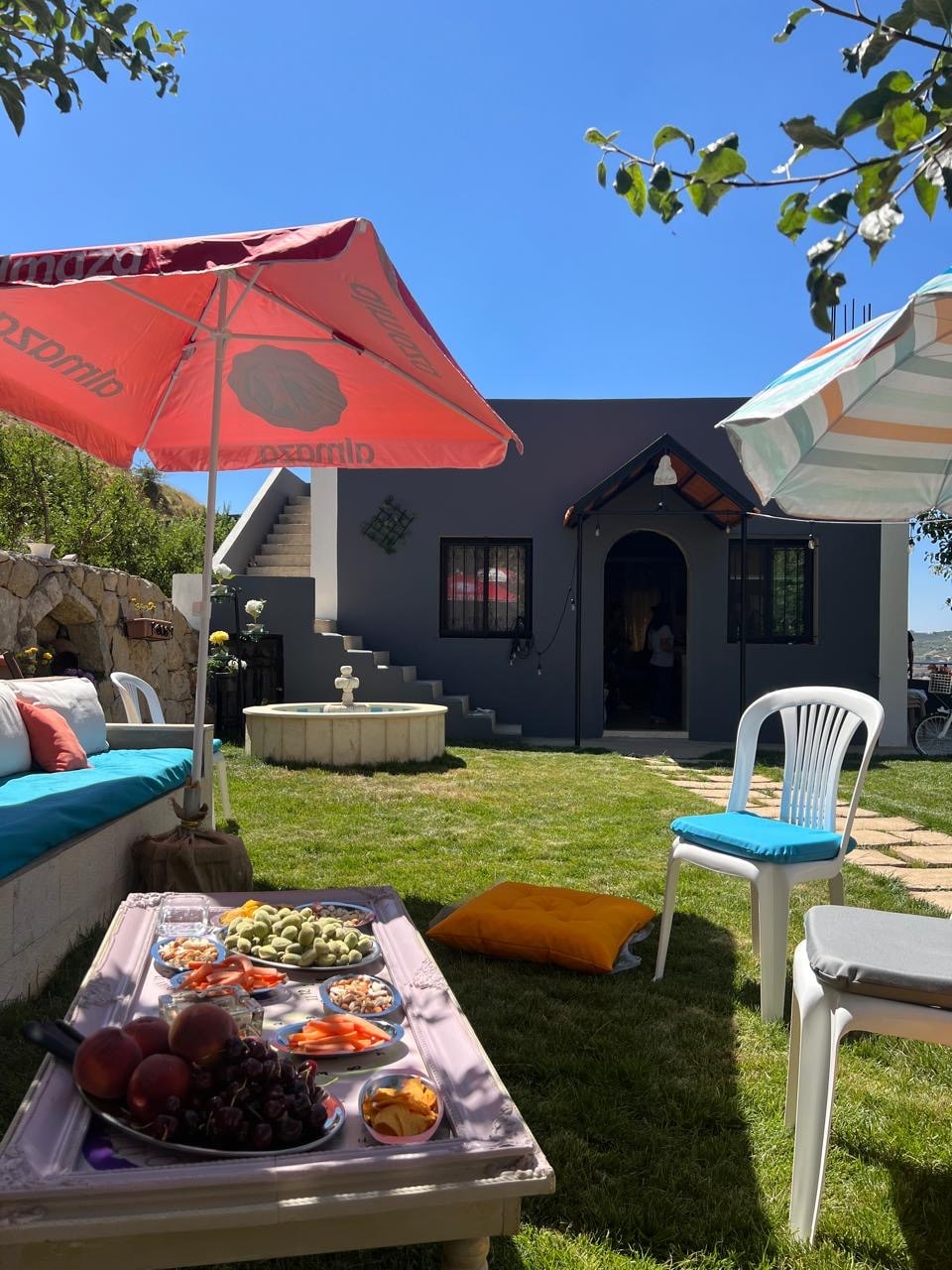 A garden area is shown with a table adorned with an assortment of fruits and snacks. Two umbrellas provide shade over comfortable seating, including a colorful cushion and a white chair. A stone staircase leads to the entrance of the house, framed by greenery.