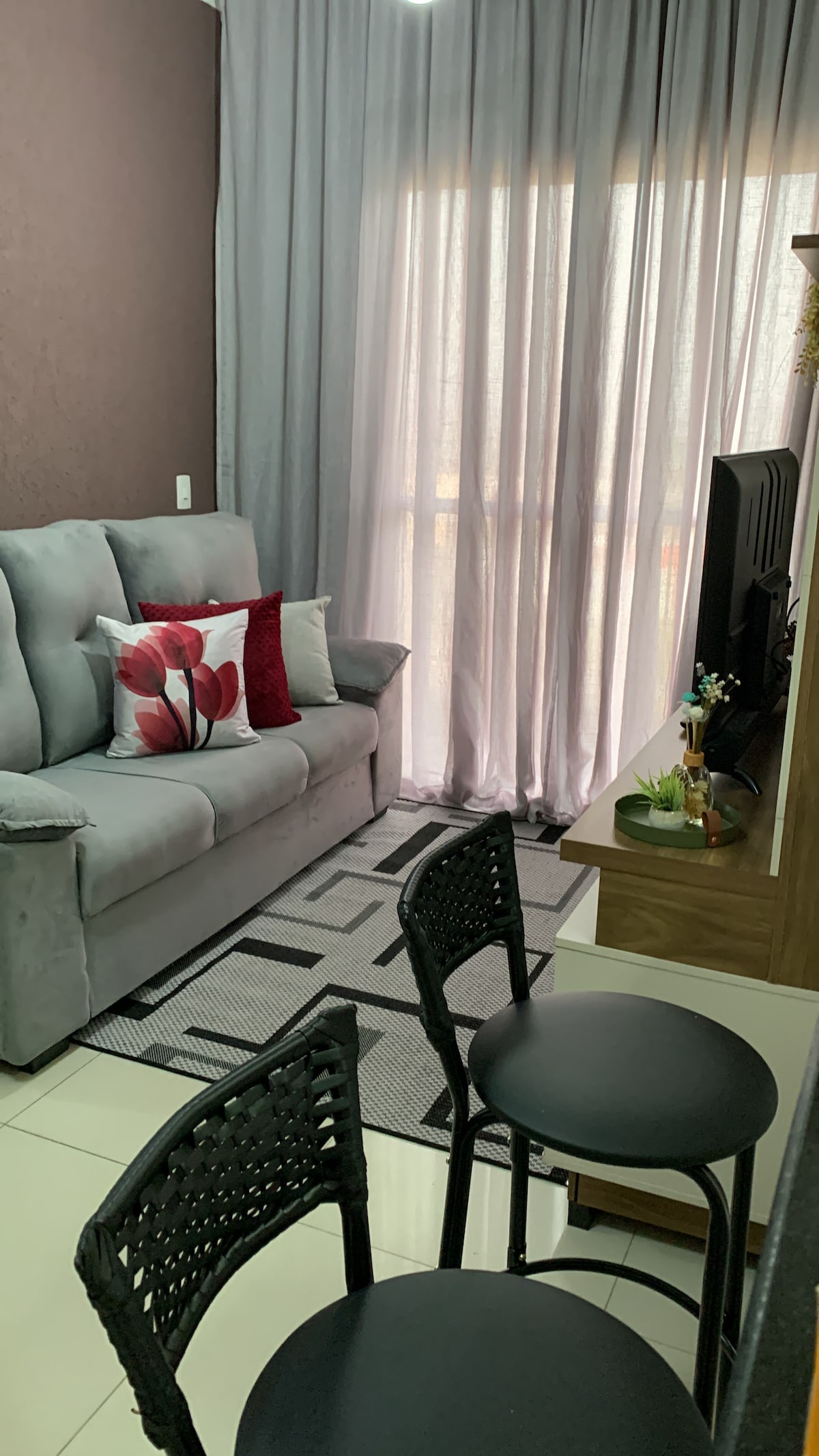 A compact living area features a soft gray sofa adorned with red and floral accent pillows. Natural light filters through sheer curtains, illuminating a patterned rug. A small dining set with black chairs is positioned adjacent to a wooden console, which displays decorative items.