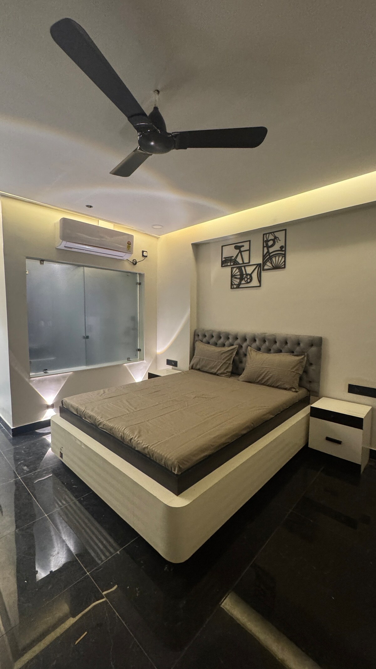 A modern bedroom features a sleek, low-profile bed with a tufted headboard and neutral bedding. A bedside table is positioned next to the bed. Above the bed, three framed artworks complement the space. A glass partition allows natural light to enter, and a ceiling fan is present.