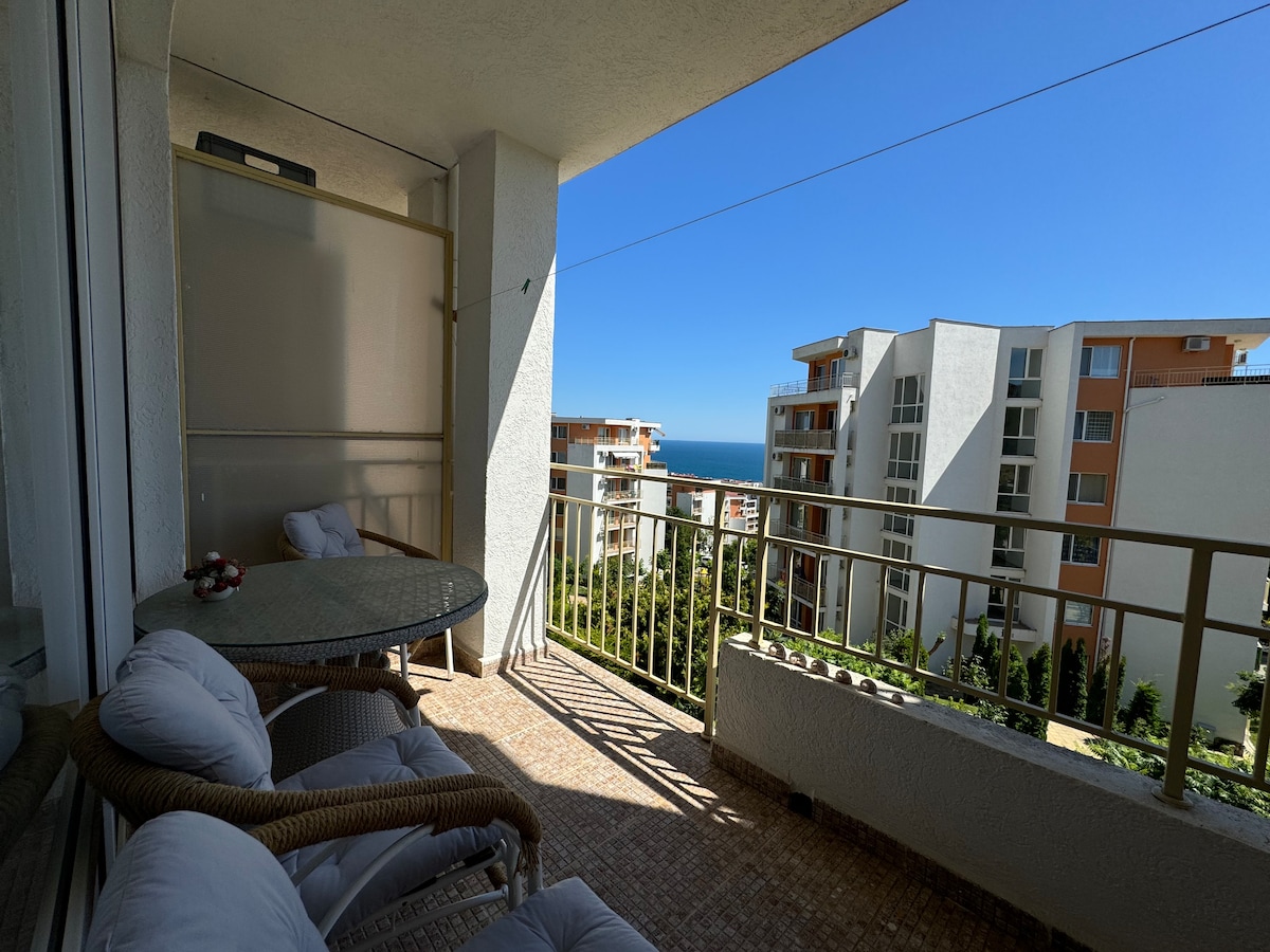 An outdoor balcony is visible, furnished with a round glass-top table and two cushioned chairs. A scenic view of the sea is presented in the background, enhanced by a clear blue sky. Lush greenery can be seen below the balcony railing.