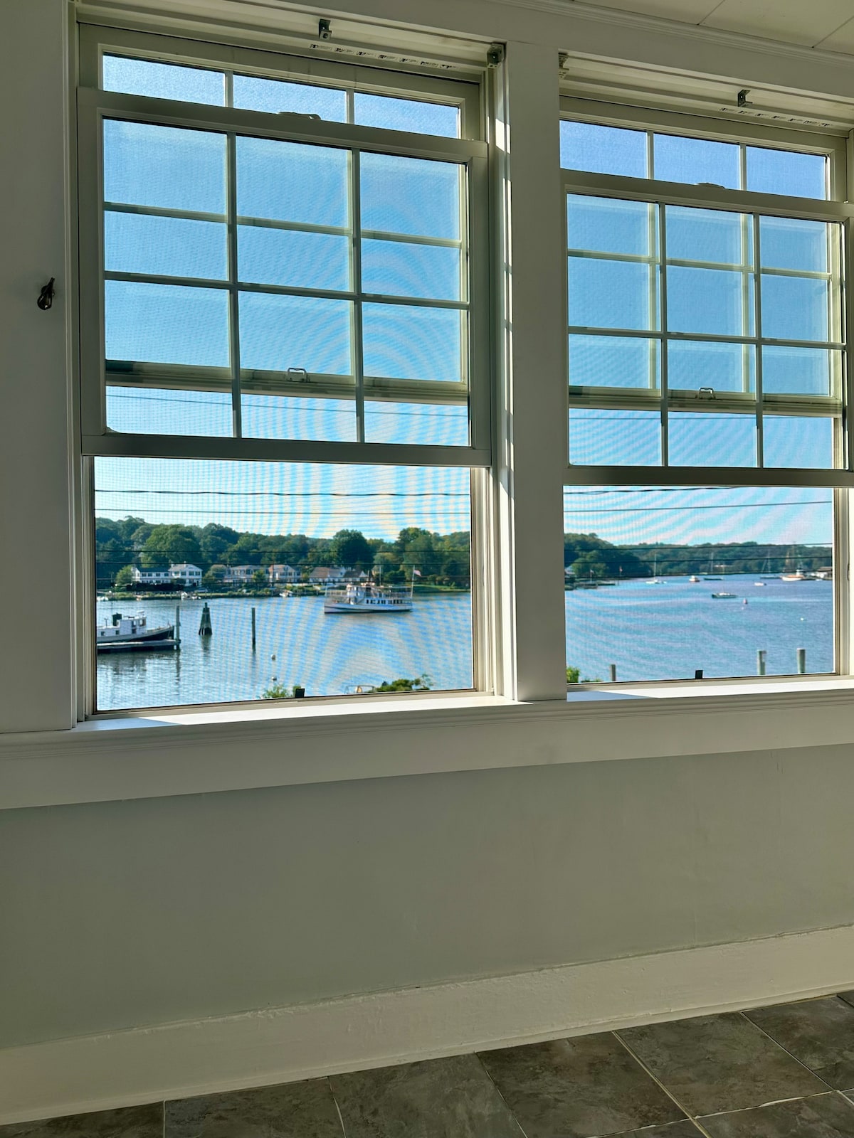 Two large windows provide unobstructed views of the river, showcasing boats on the water and lush greenery along the shore. Natural light fills the space, enhancing the serene ambiance of this historic apartment.