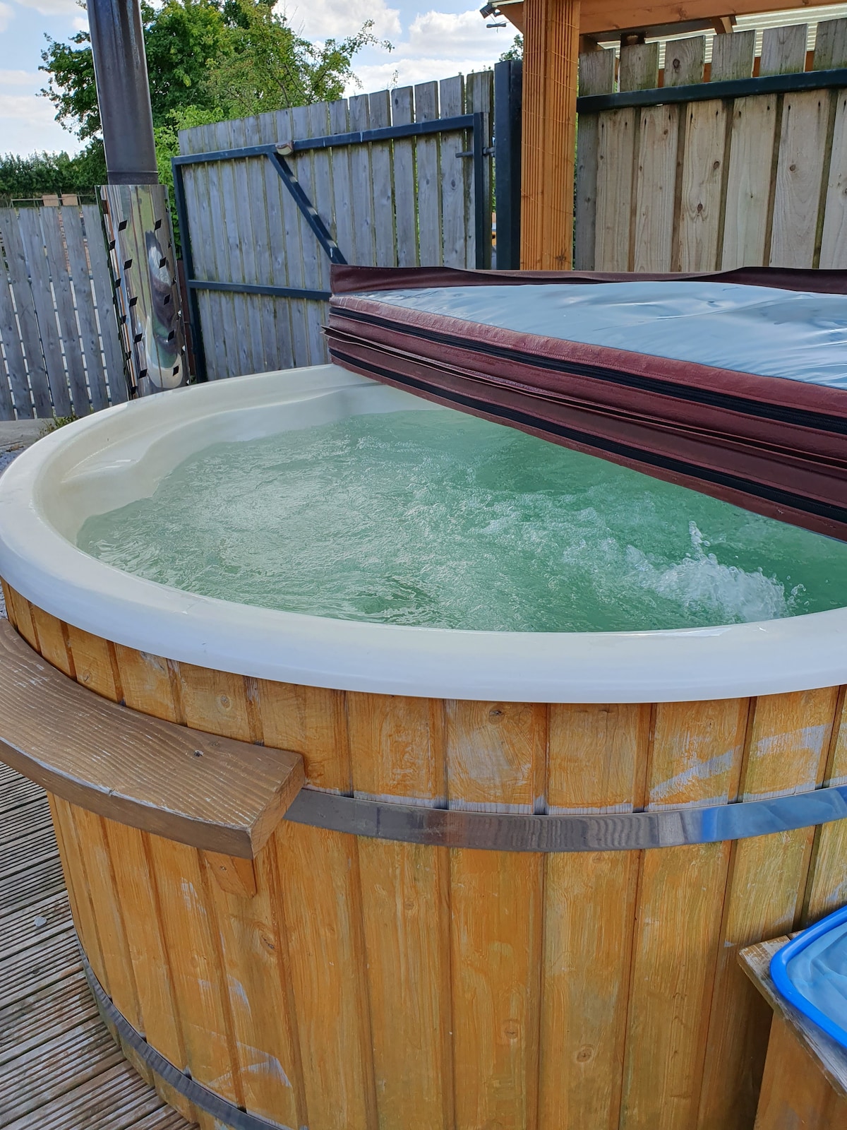 A wooden hot tub filled with bubbling water is shown, surrounded by a deck. A protective cover rests partially open on the tub, allowing for easy access. The enclosed area features wooden fencing, providing a sense of privacy in the outdoor setting.