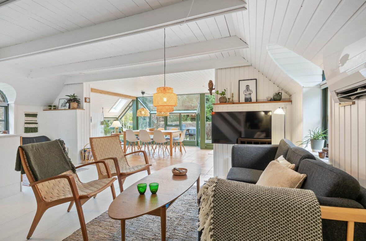 A spacious living area is highlighted by wooden beams and large windows offering natural light. Comfortable seating includes a dark sofa and woven chairs, complemented by a coffee table. A dining space with a wooden table and multiple chairs is visible in the background, with greenery framing the view.