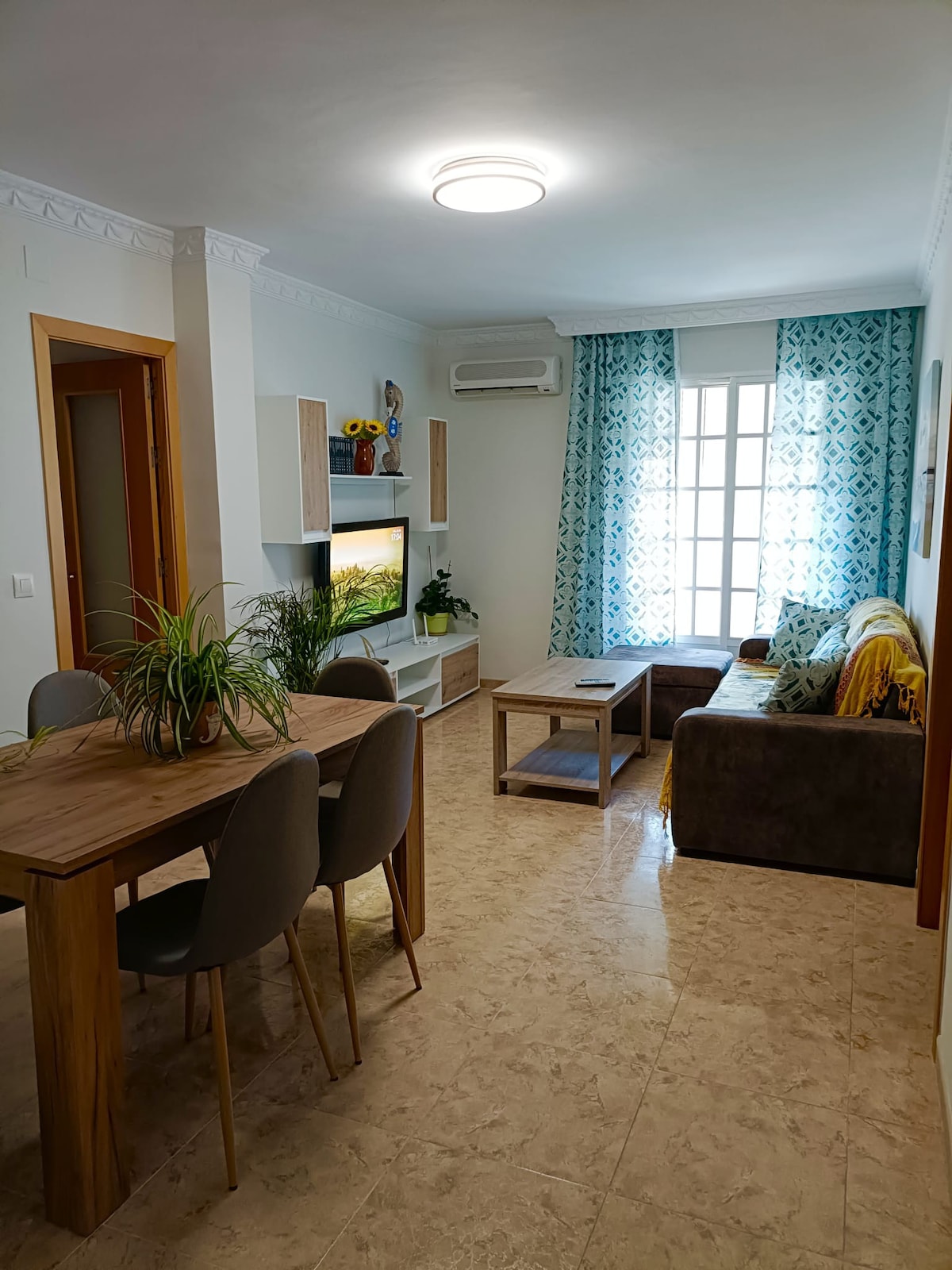 A welcoming living area features a comfortable sofa adorned with a throw, a wooden coffee table, and a dining table with rounded chairs. Natural light enters through a window, accentuated by light blue curtains, while a television is positioned on a wall-mounted unit.