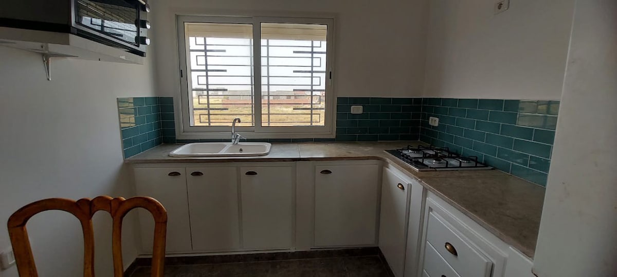 The kitchen features a functional layout with white cabinetry and teal backsplash tiles. A gas stove is positioned on the right side, while a double sink is situated under a large window that allows natural light to enter the space.